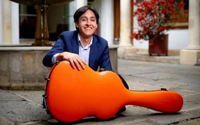 Artista sonriendo, usando un traje azul, sentado en el suelo, frente al estuche naranja de una guitarra