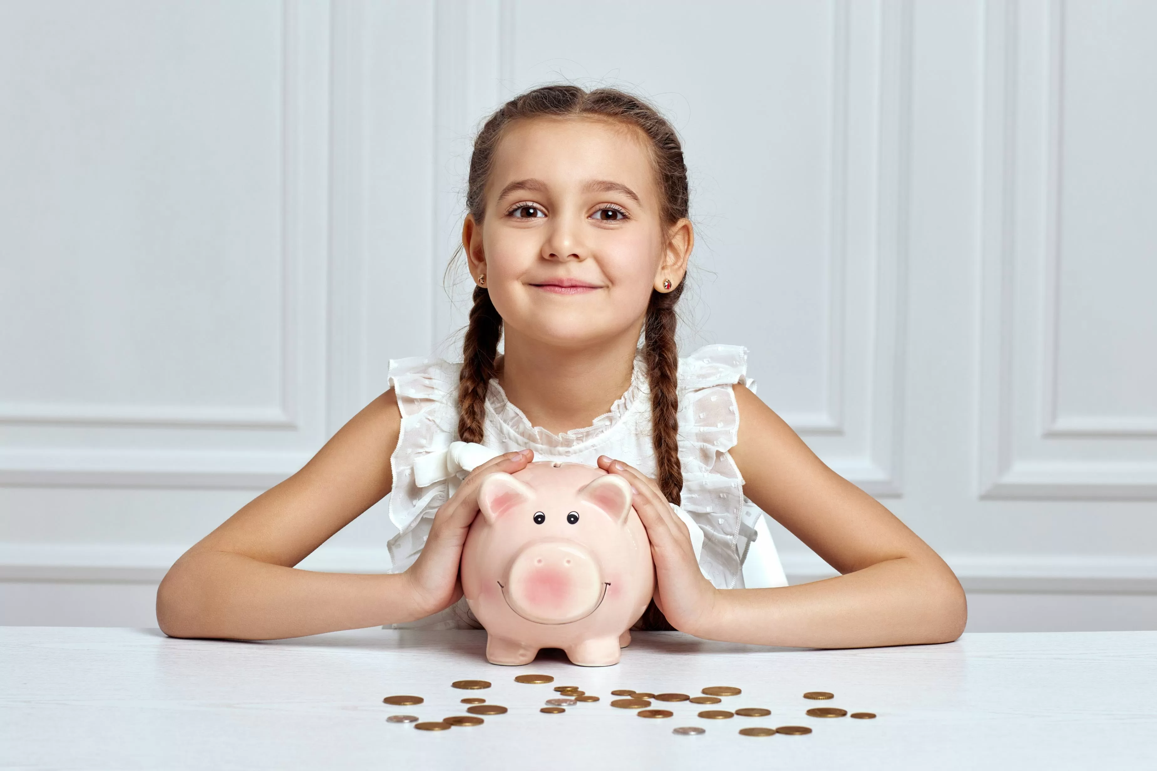 Una niña de trenzas con una alcancía de marranito y algunas monedas sobre una mesa blanca