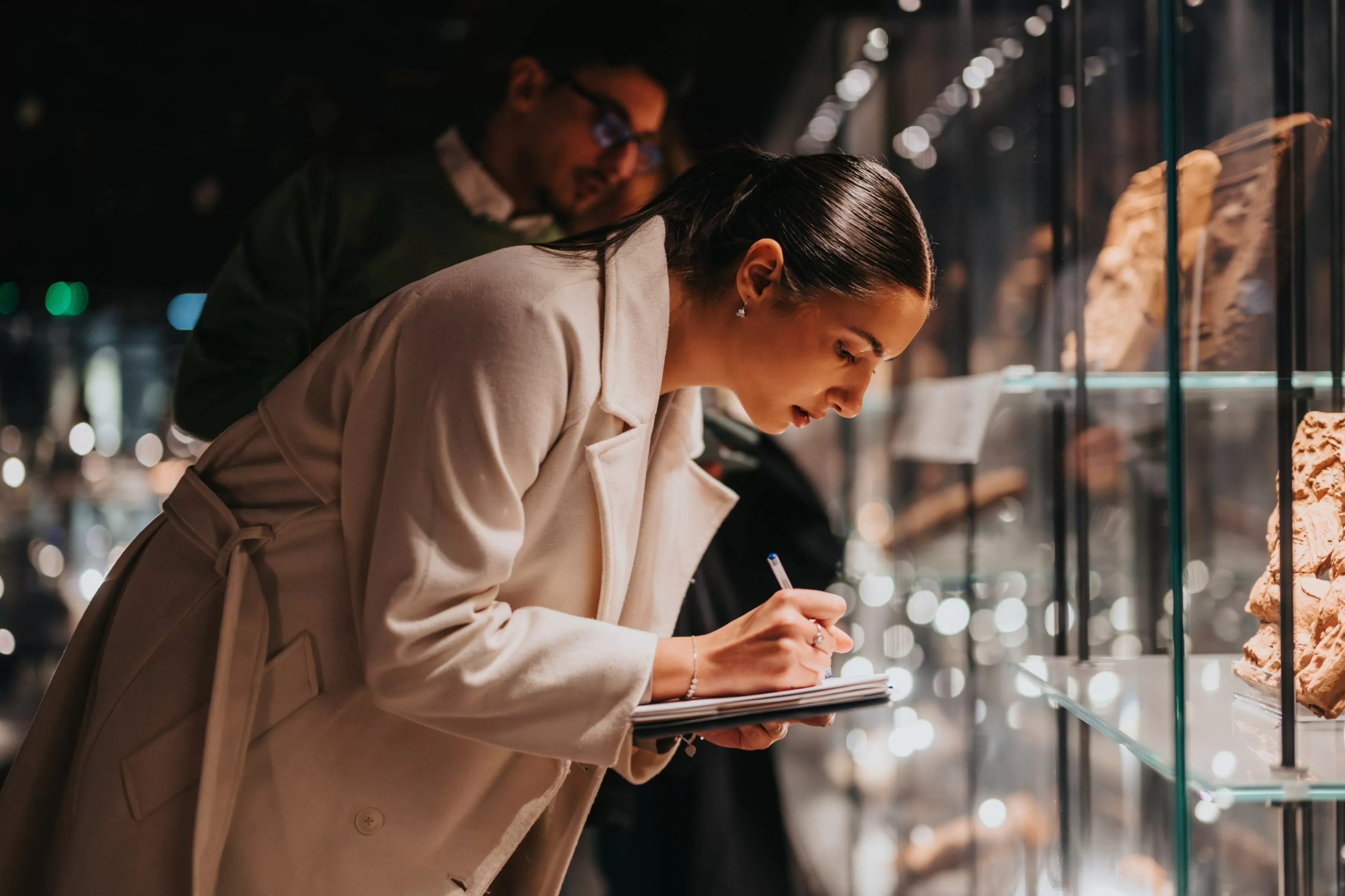 Una mujer observa y toma notas en una exposición del museo, mostrando artefactos en vitrinas iluminadas,