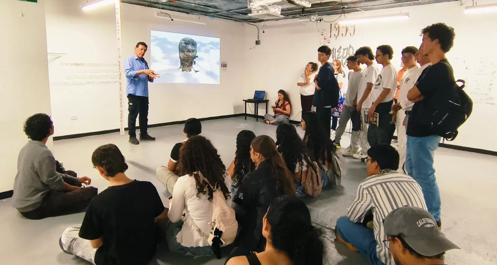 La imagen muestra a un grupo de personas escuchando a un expositor de una obra