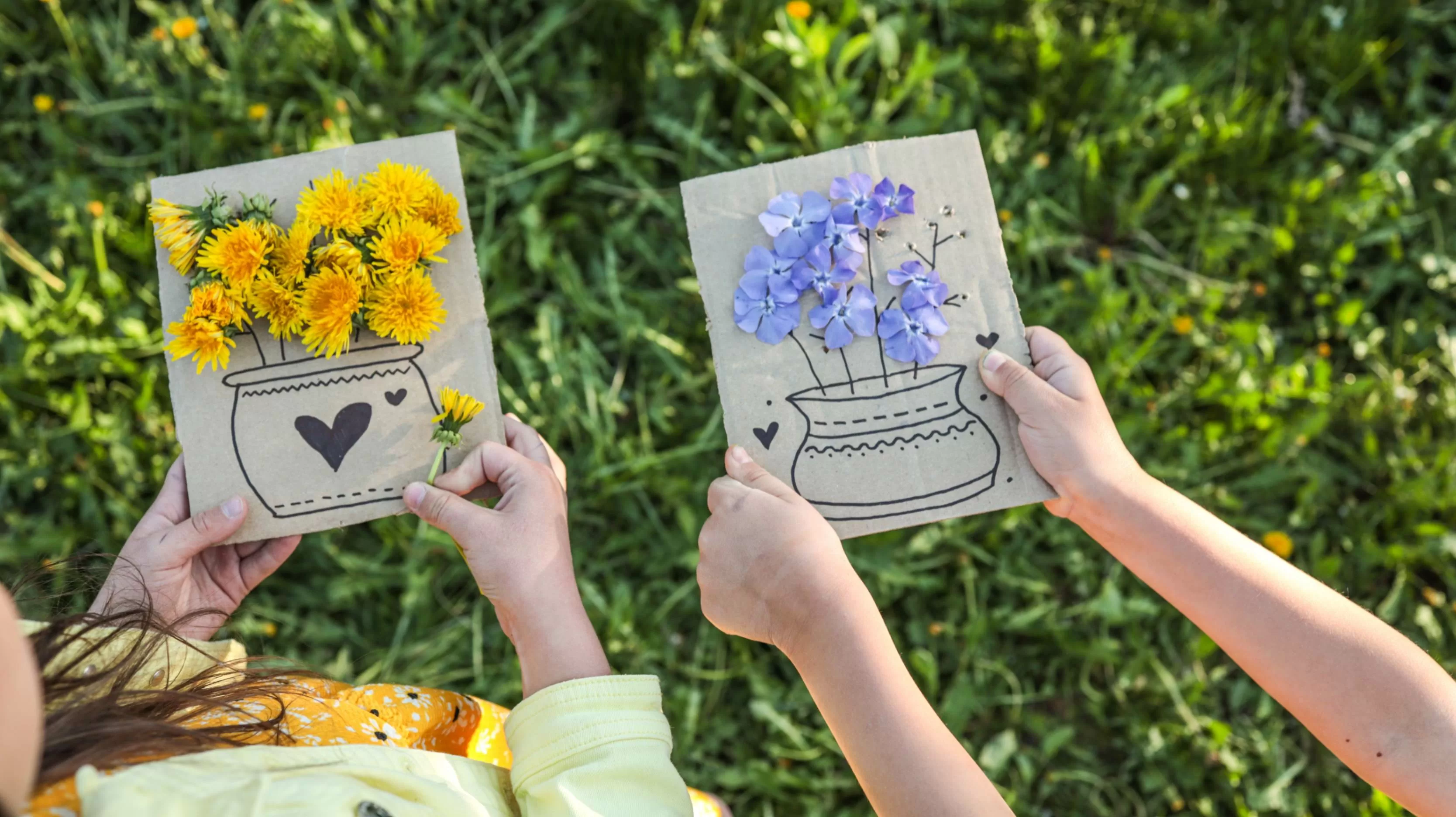 Dos niñas sostienen tarjetas hechas a mano con jarrones dibujados llenos de flores amarillas y azules reales sobre un fondo de pasto