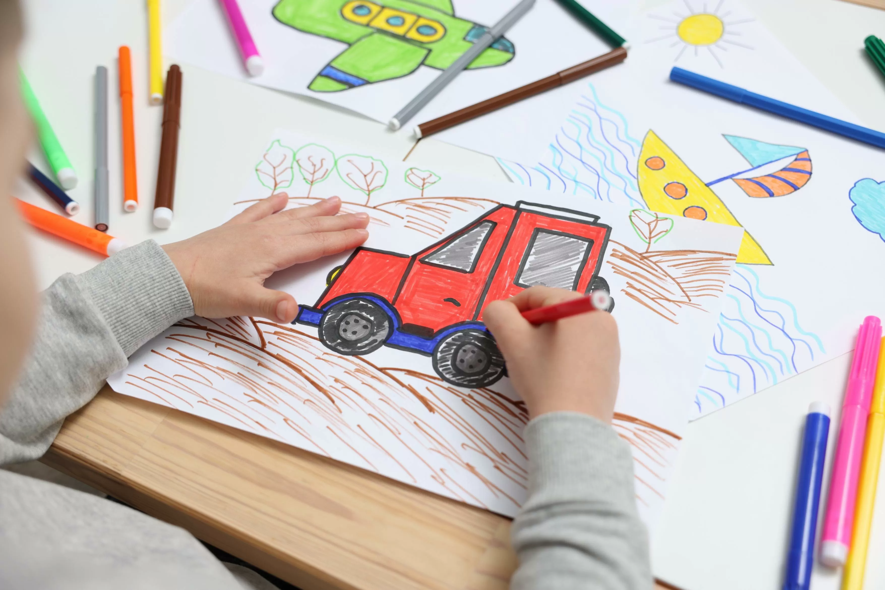 Niño pequeño que dibuja un carro rojo con marcadores en la mesa, primer plano