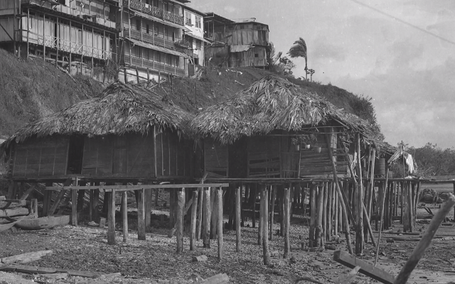 Fotografía en blanco y negro, que enseña una casa en Buenaventura, un modelo de vivienda palafítica, con sus grandes bases incrustadas en la arena, elevando la casa sobre el mar